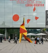 Stahlskulptur auf einem dreibeinigen Rot, gelben Gestell mit einem beweglichen Stabelement an dem sich auf der einen Seite ein großer Kreis und auf der anderen Seite mehrere kleine rautenförmige Elemente befinden in kräftigen Rot. Weitere Informationen im Text.