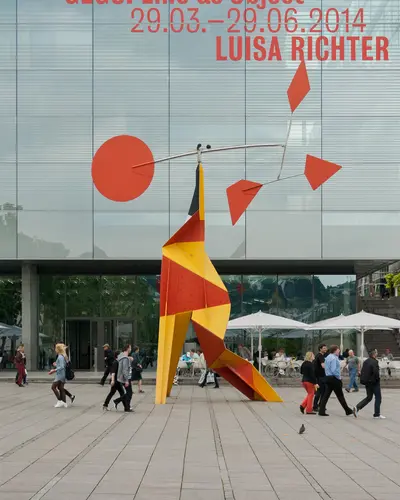 Stahlskulptur auf einem dreibeinigen Rot, gelben Gestell mit einem beweglichen Stabelement an dem sich auf der einen Seite ein großer Kreis und auf der anderen Seite mehrere kleine rautenförmige Elemente befinden in kräftigen Rot. Weitere Informationen im Text.