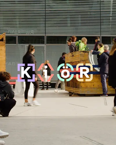 KiöR-Logo auf einem Hintergrund, der die Installation Platzprobe auf dem kleinen Schlossplatz zeigt.